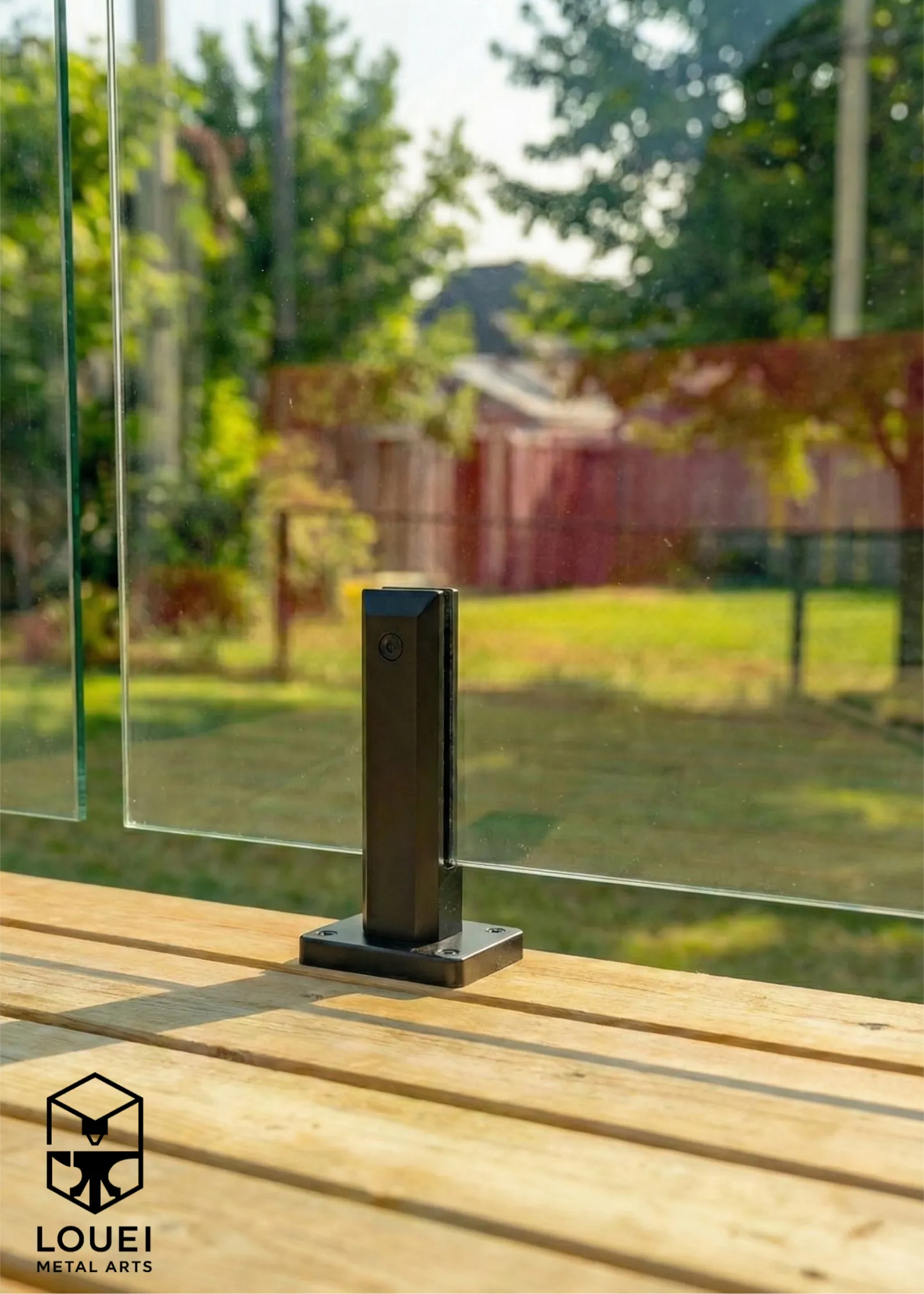 Close-up of a black railing post with base plate mounted to a wood deck, supporting an exterior glass railing panel.