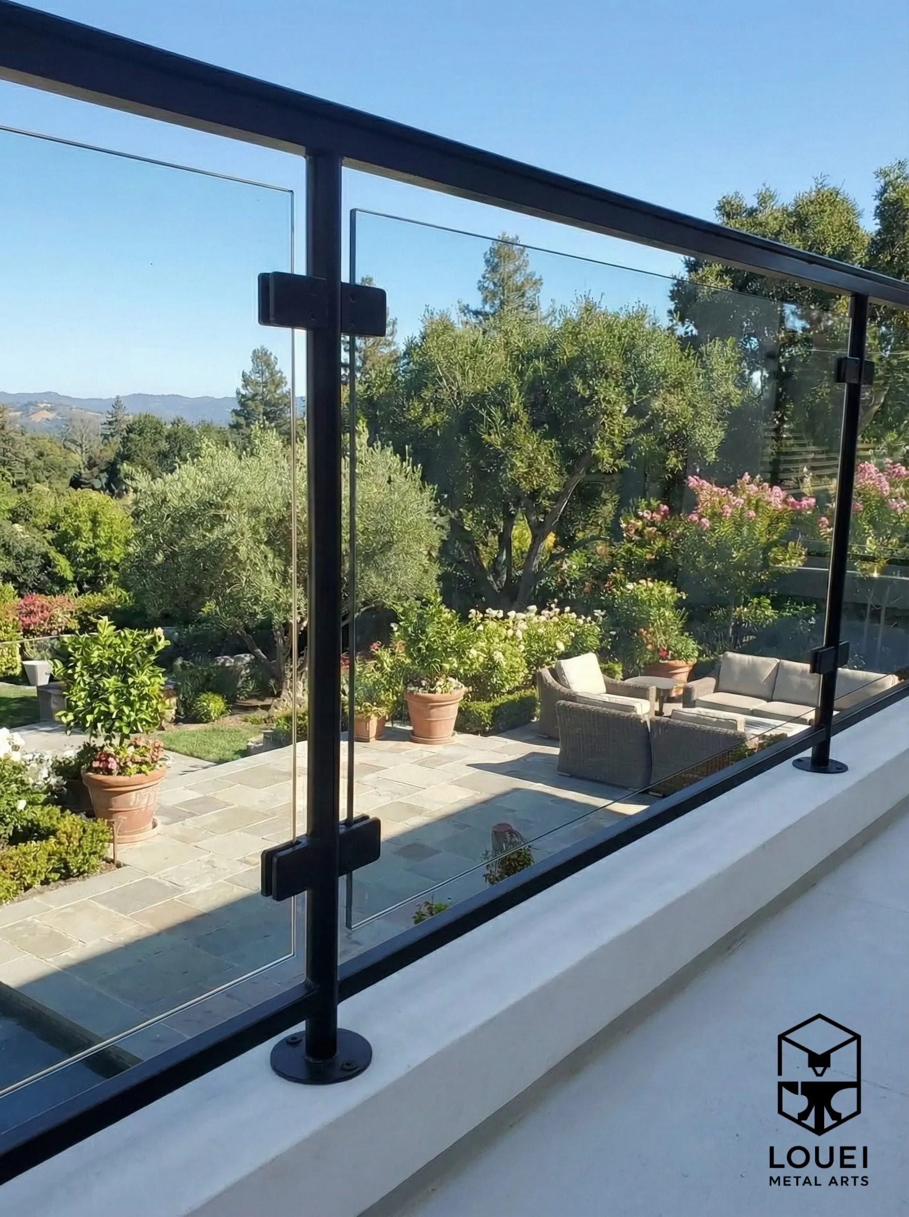Exterior glass railing with black posts and a black top rail mounted on a terrace edge overlooking a landscaped backyard.
