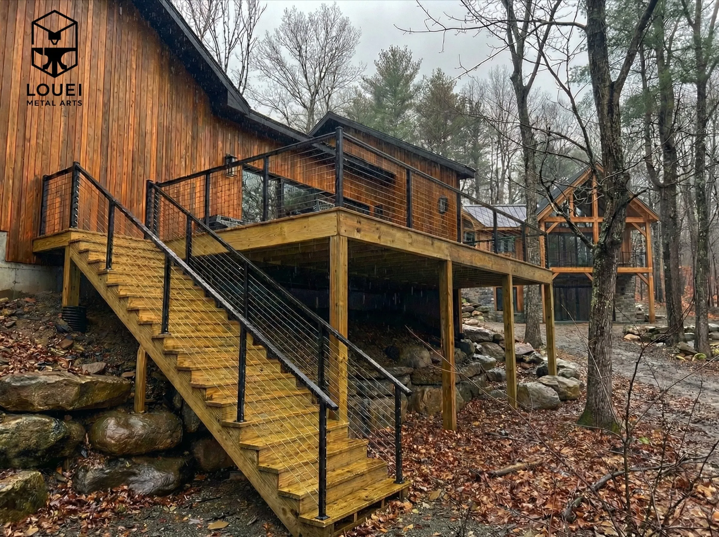 Elevated wood deck with black metal railing and wire mesh infill on outdoor stairs beside a cedar cabin in the woods.