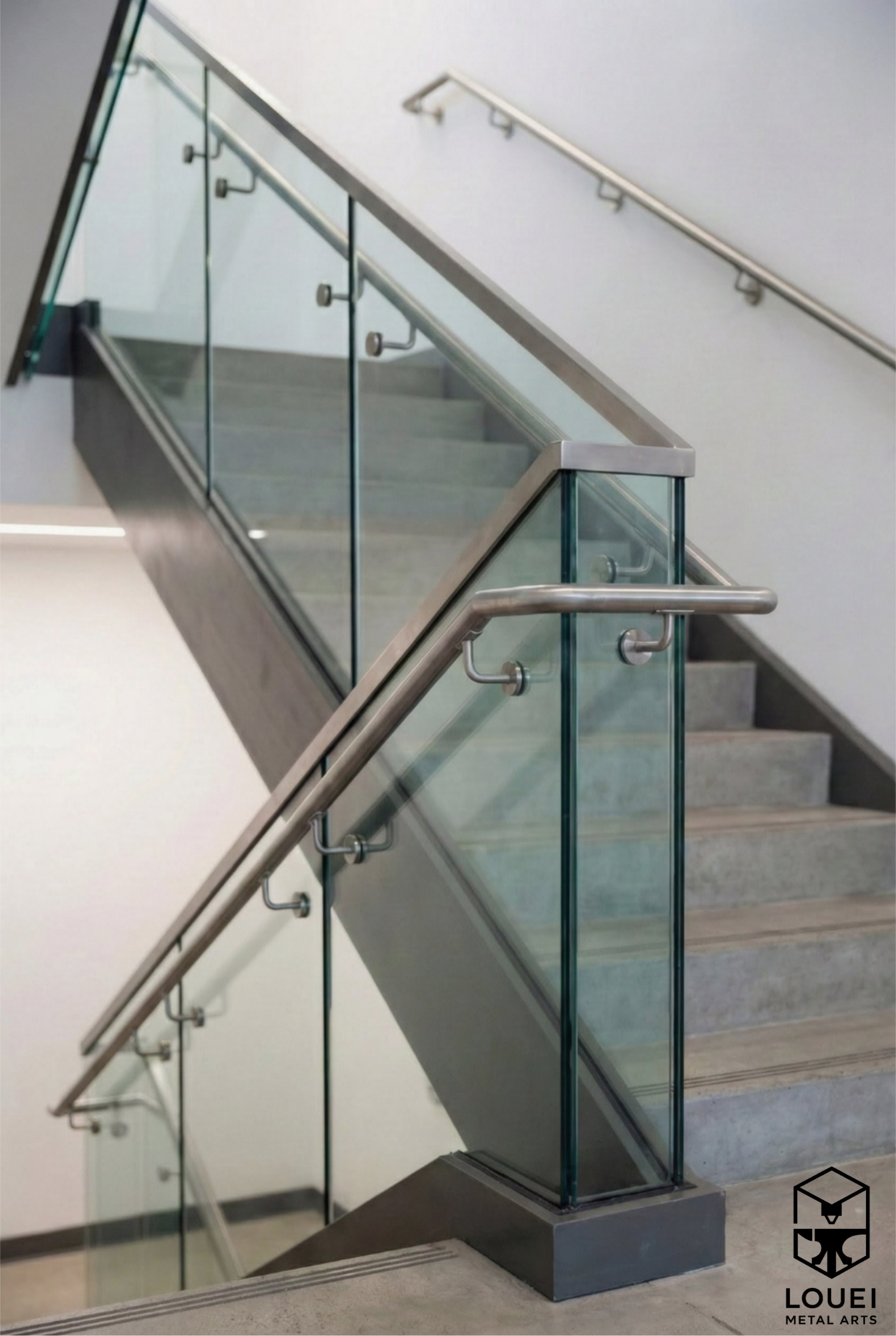 Stainless steel handrail mounted to a glass stair guardrail alongside a concrete staircase with a metal cap rail.