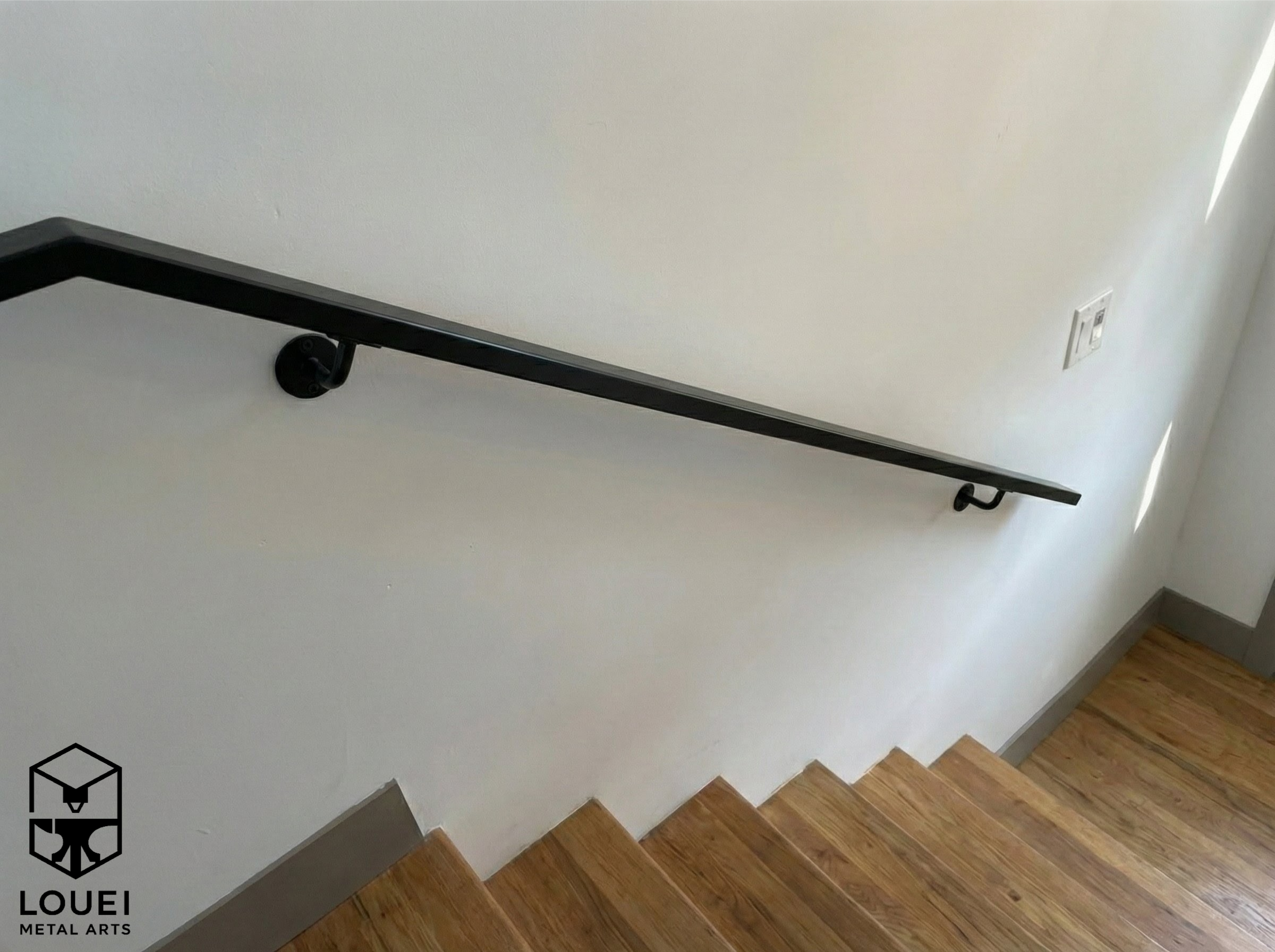 Wall-mounted black square steel handrail with metal brackets installed along an interior wood staircase.