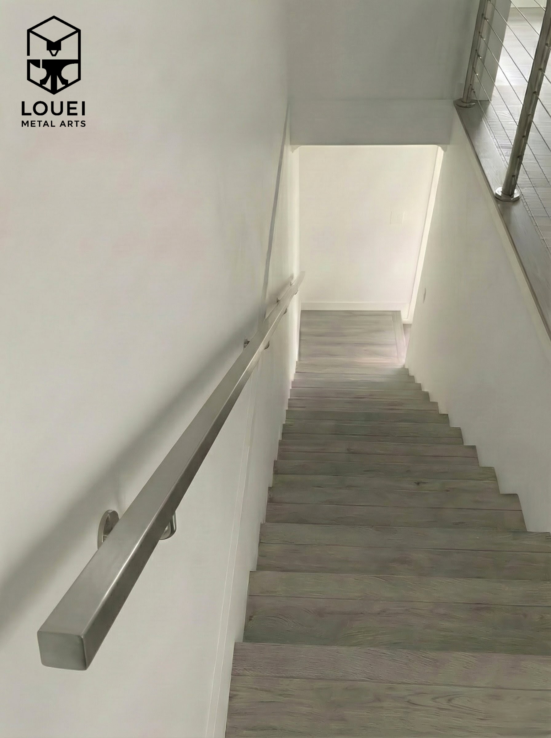 Wall-mounted stainless steel square handrail installed along an interior staircase with light wood treads.