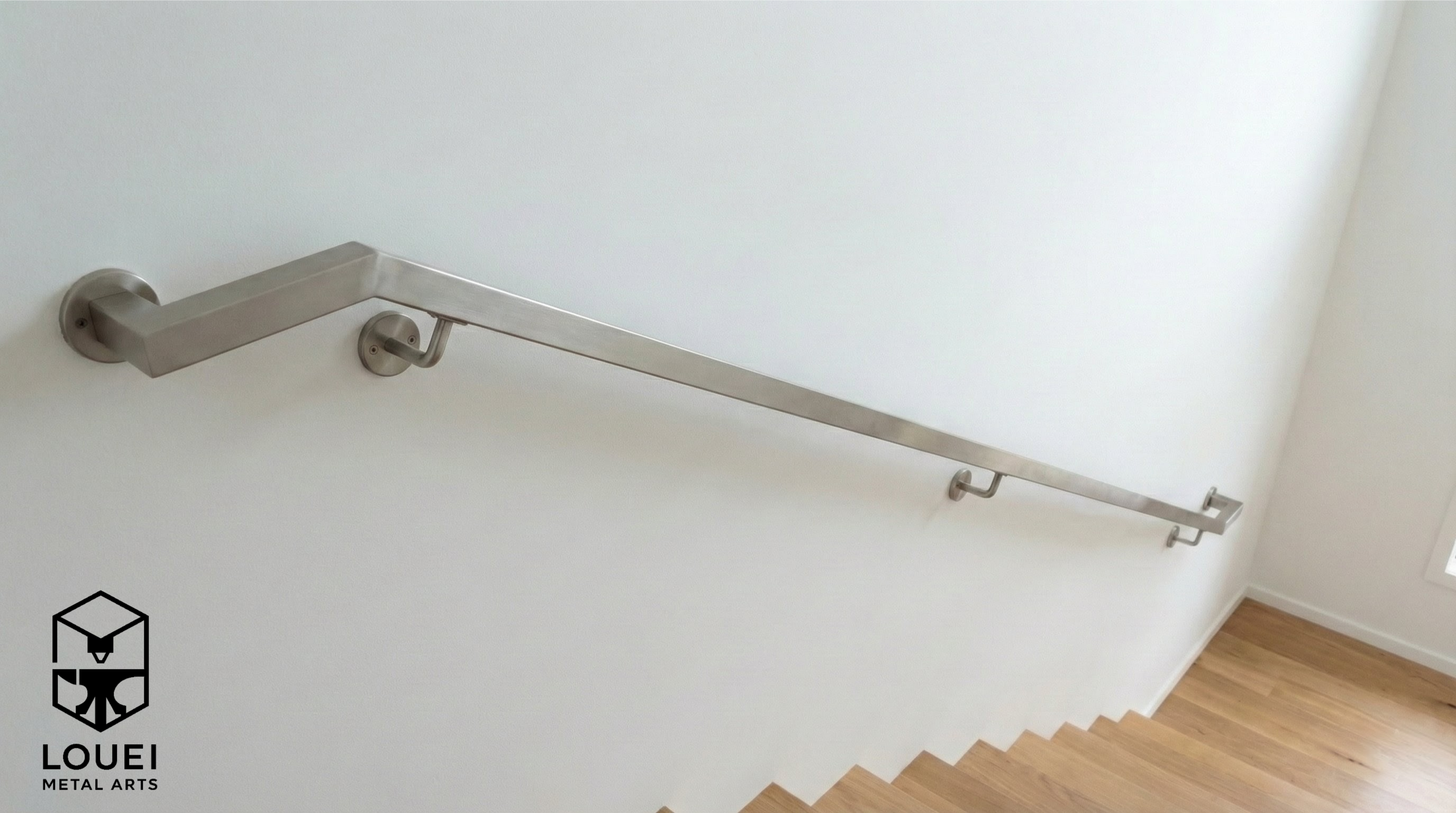 Wall-mounted stainless steel square handrail with return end, installed on an interior stair wall above wood flooring.