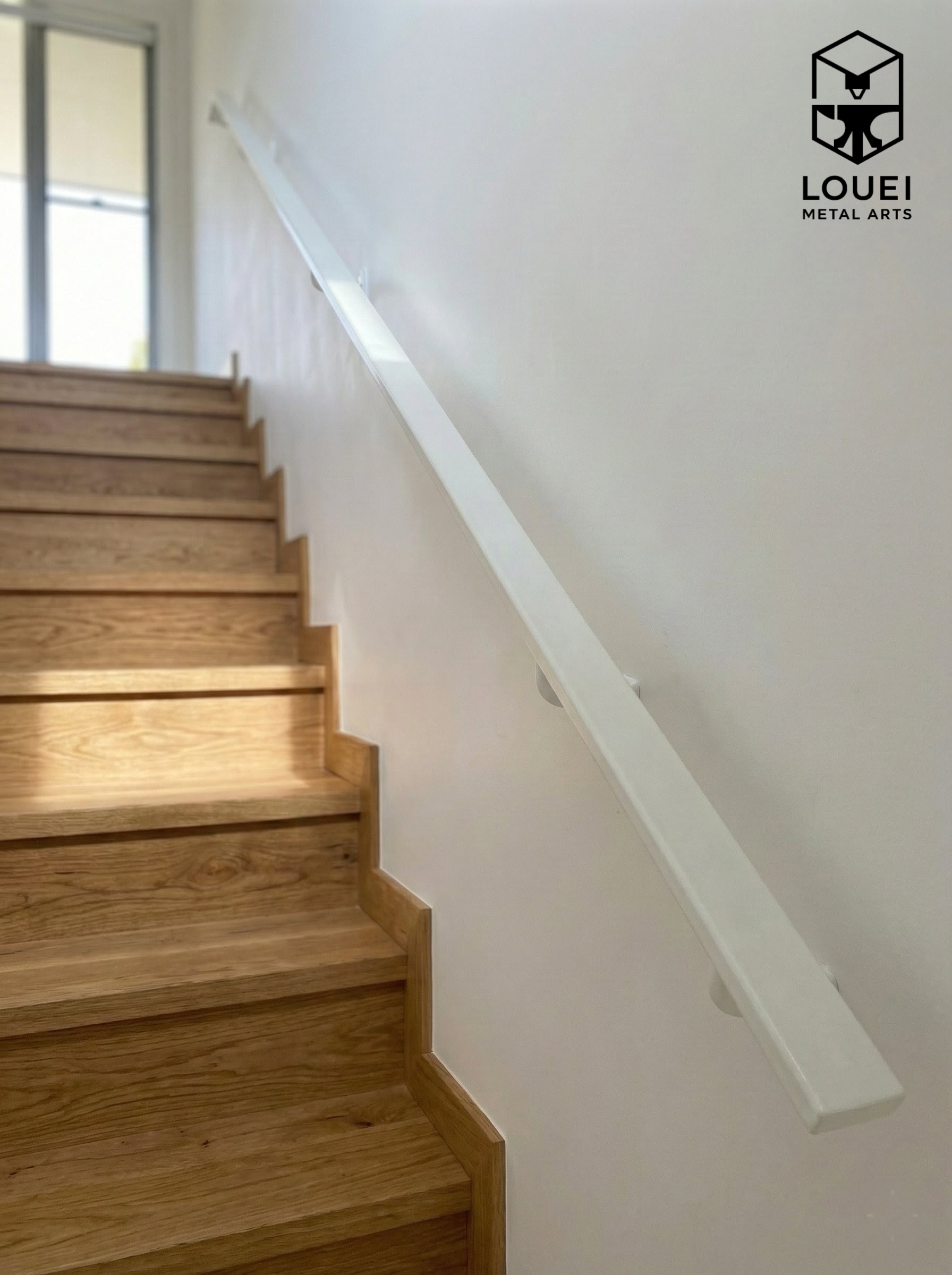 Minimalist wall-mounted white handrail installed along an interior wood staircase.